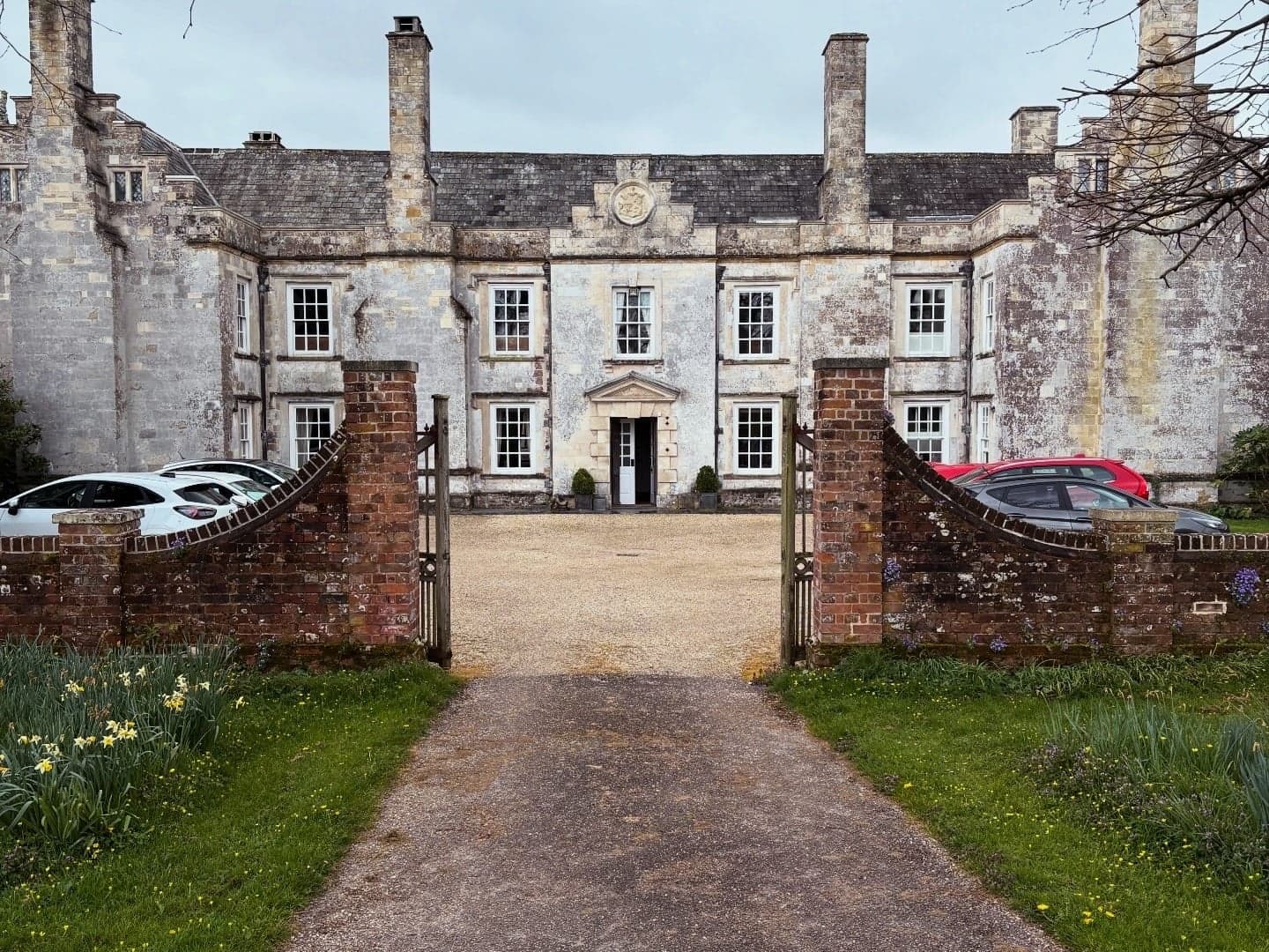 The manor house entrance seen past parked cars and iron gates.